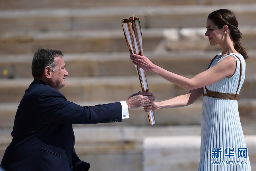 希臘奧委會主席斯皮羅斯·卡普拉洛斯（左）從扮演最高女祭司的希臘女演員桑西·喬治烏手中接過聖火火炬。  新華社發（阿裡斯·麥西尼攝）